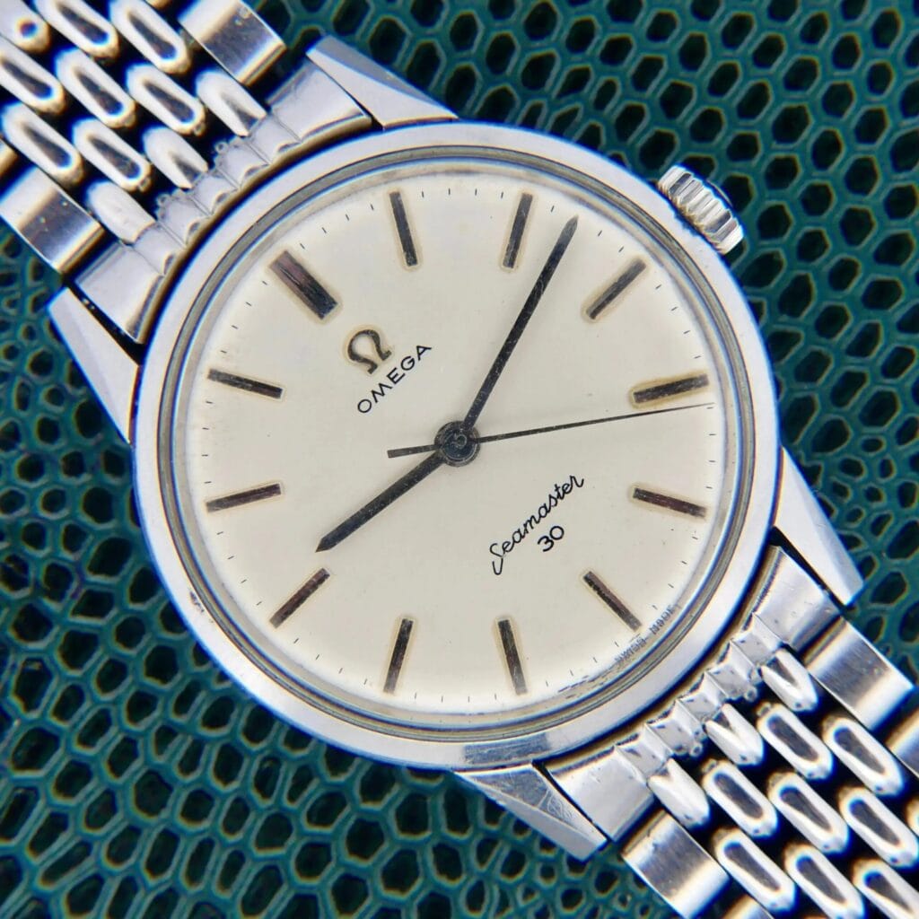 A close-up of a vintage Omega Seamaster 30 Cal. 286 Reference 135003 watch with a silver band and white dial on a textured surface.