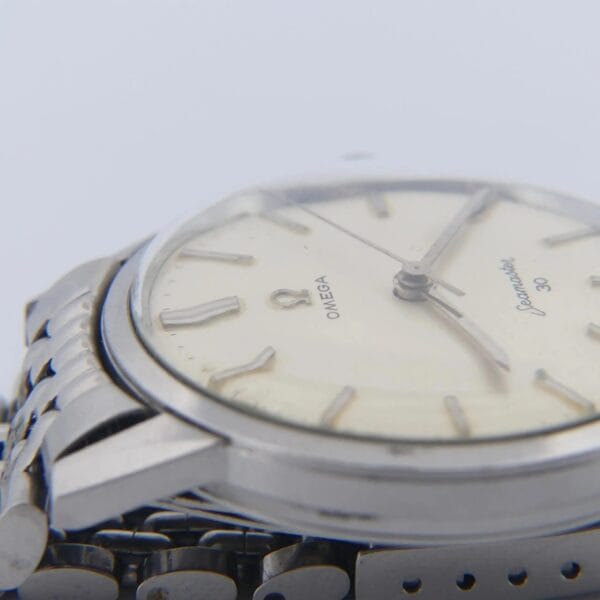 Close-up of an Omega Seamaster 30 Cal. 286 Reference 135003, featuring a stainless steel case and bracelet. The watch has a white dial with simple hour markers and hands, highlighting the brand name and logo. Focus is on the side of the watch face and part of the metal link band.