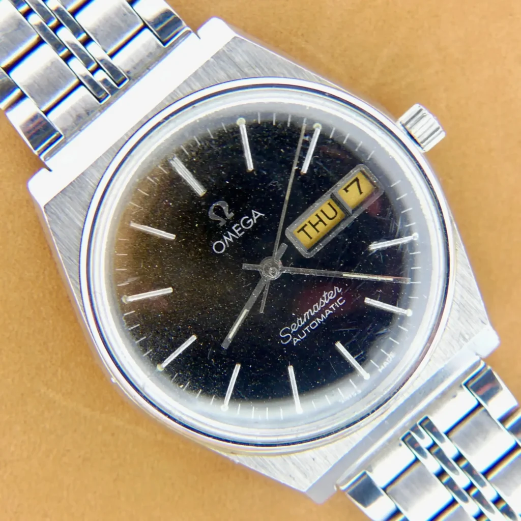 A close-up of an Omega Seamaster Automatic Ref 166.210 Tropic Dial wristwatch with a silver metal band, black dial, and day-date window showing THU 7, placed on a tan surface.