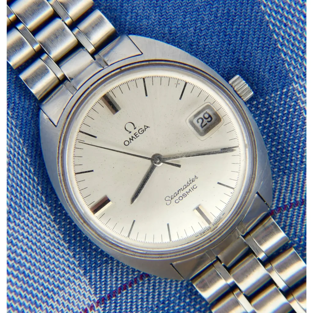 An Omega Seamaster Cosmic Ref. 136.017 Caliber 613 wristwatch with a metal band is displayed on blue fabric, featuring a white dial, silver hour markers and hands, and the date set to 29.