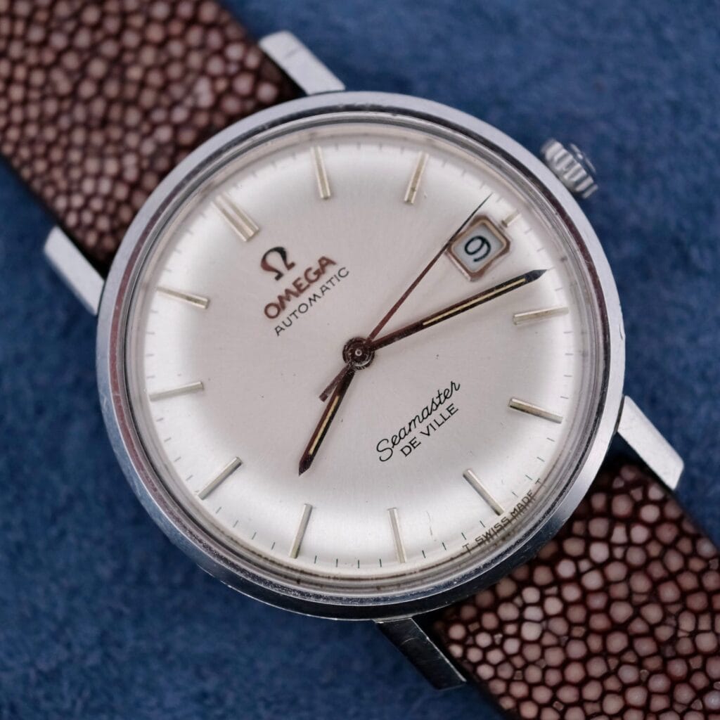 A close-up of an Omega Seamaster Deville Automatic Cal. 560 wristwatch featuring a silver dial, date window at 3 oclock, and brown textured leather strap, displayed on a blue surface.