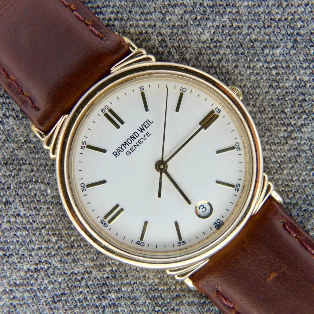 A Raymond Weil 5531 Men’s Swiss Quartz Watch with a gold tone case rests on a gray textured surface, highlighting its timeless date feature and classic craftsmanship.