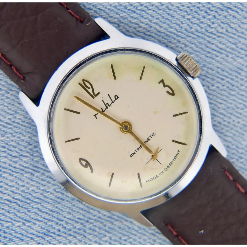 A close-up of the Rhula UMF Germany Cream Dial round wristwatch with a brown leather strap and gold hour markers, set on light denim, shows the time as about 11:02.