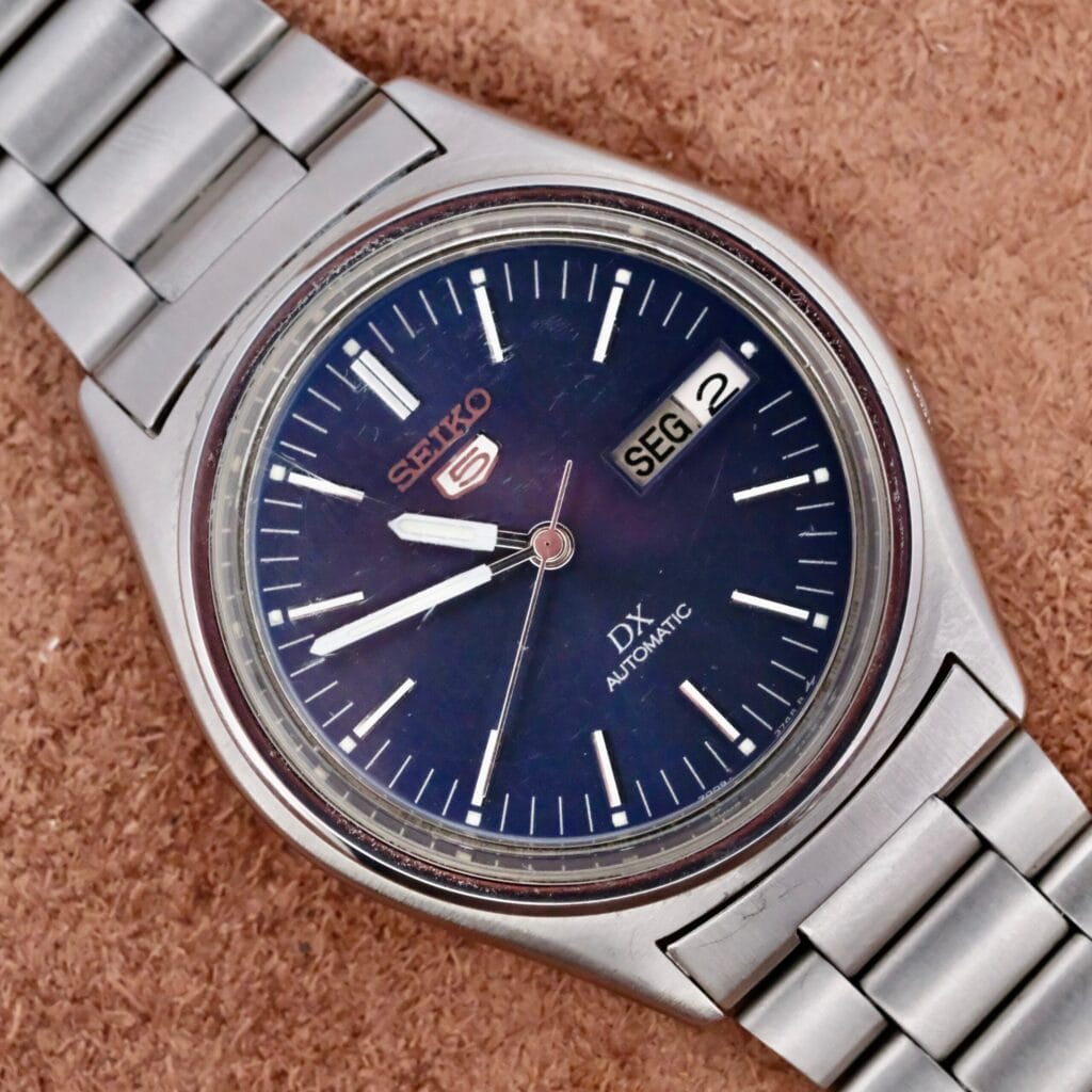 A Seiko Automatic Day/Date Cal. 7009 wristwatch with a dark blue dial, silver hour markers, day-date window showing SEG 2, and a metal band is displayed on a brown textured surface.