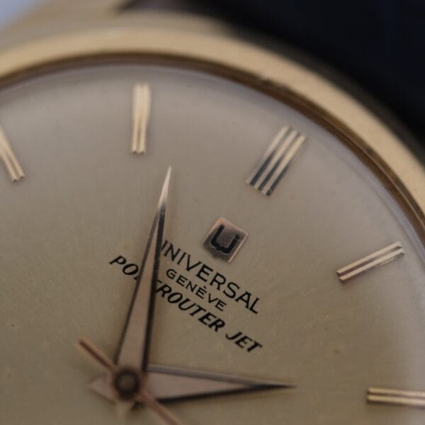 Close-up of a Universal Geneve Polerouter Jet 18k Cal. 215-9 wristwatch, showing gold hour markers, thin black minute markers, and black text on a brushed metal dial with the hands near the center.