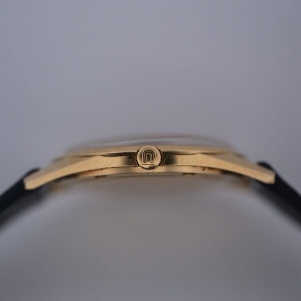 Close-up side view of a Universal Geneve Polerouter Jet 18k Cal. 215-9 gold case with a crown featuring an engraved logo. The watch has a black strap on both ends, displayed against a plain light background.