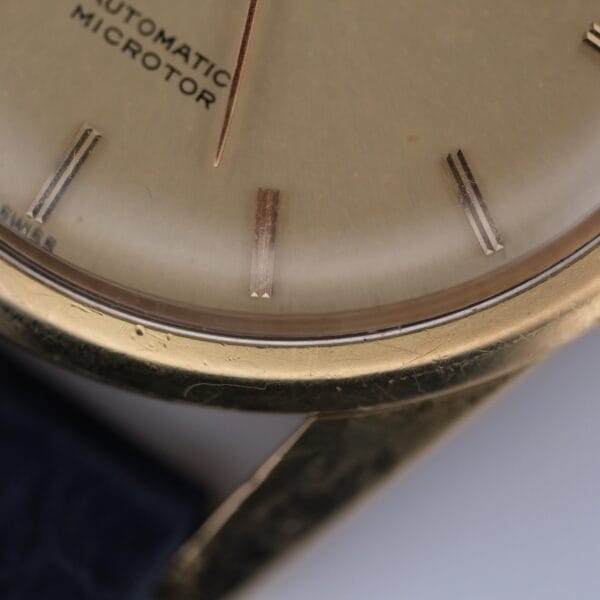 Close-up of a Universal Geneve Polerouter Jet 18k Cal. 215-9 watch face with gold-toned casing, showing the edge of the dial, part of the crystal, and metallic hour markers; AUTOMATIC MICROTOR text is partially visible.