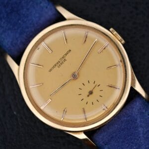 A Vacheron Constantin V453 Calatrava 18k Gold wristwatch (1950s, 34mm) featuring a round beige dial, thin hour markers, blue strap, and small seconds subdial at 6 o’clock, displayed on a black surface.