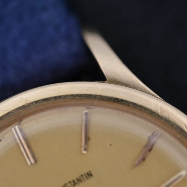 Close-up of a Vacheron Constantin V453 Calatrava 18k Gold 1950s 34mm VC wristwatch, showing part of the gold face with gold hour markers and bezel edge. The blurred dark and blue background highlights the partially visible brand near the bottom.