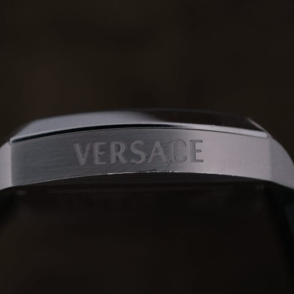 A close-up of the Versace Character Tonneau Swiss Made Chronograph showcases the engraved VERSACE logo on its metallic surface against a dark background.