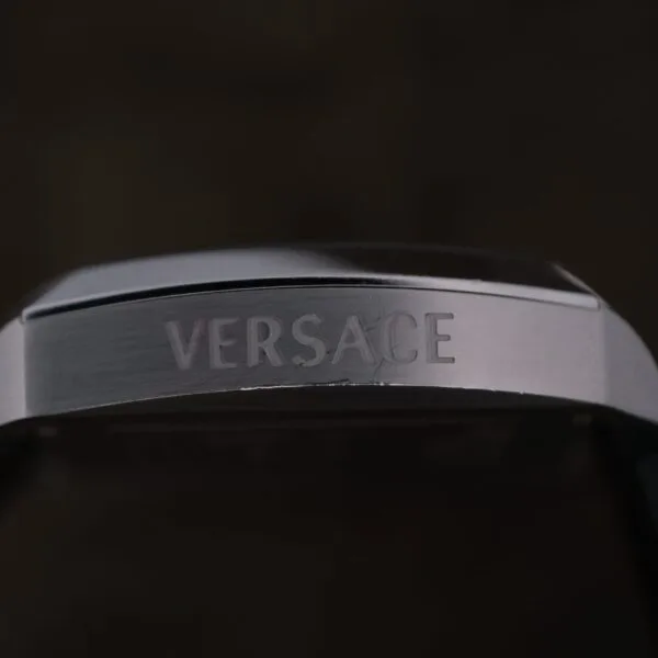 A close-up of the Versace Character Tonneau Swiss Made Chronograph showcases the engraved VERSACE logo on its metallic surface against a dark background.