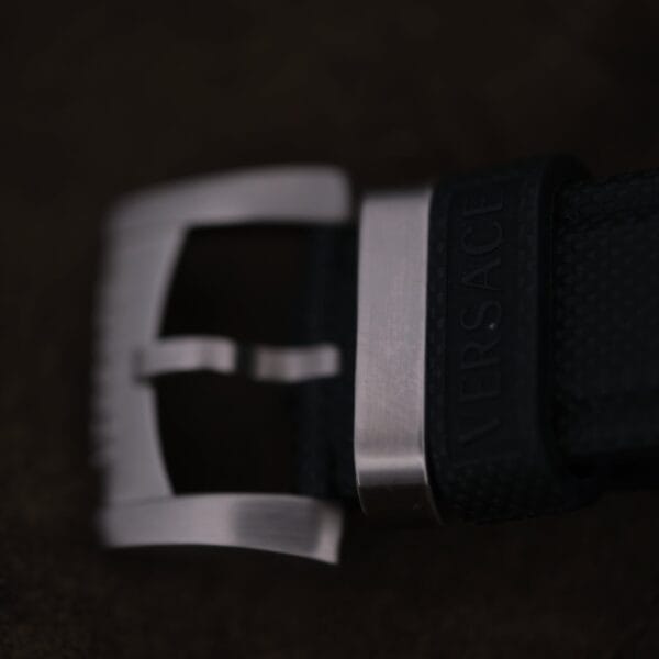 Close-up of the Versace Character Tonneau Swiss Made Chronograph’s brushed metal buckle on a textured black strap embossed with Versace, set against a dark, blurred background.
