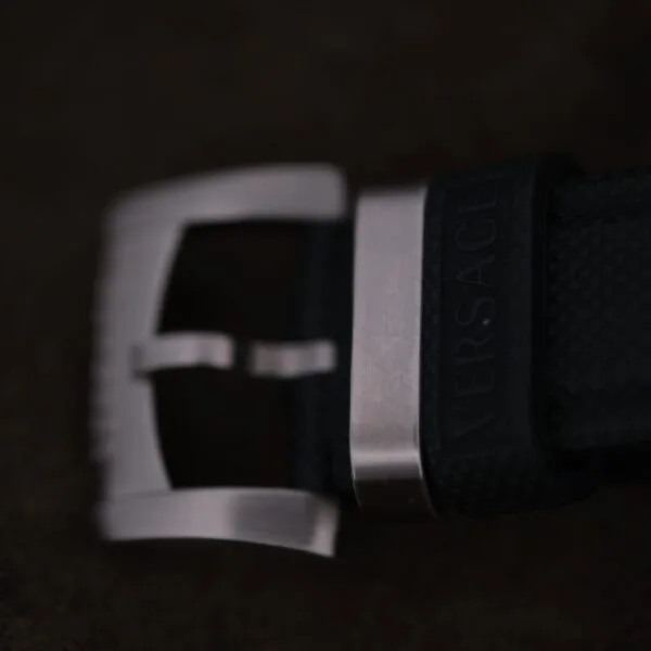 Close-up of the Versace Character Tonneau Swiss Made Chronograph’s brushed metal buckle on a textured black strap embossed with Versace, set against a dark, blurred background.