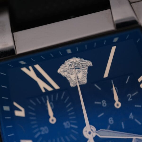 Close-up of the Versace Character Tonneau Swiss Made Chronograph, showcasing a blue dial with silver hands, Roman numerals, and a striking Medusa head logo at the top center. Parts of its metal band are also visible.