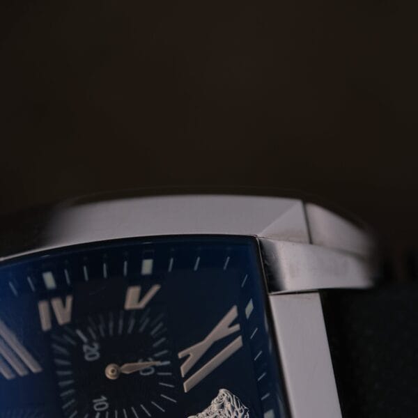Close-up of the Versace Character Tonneau Swiss Made Chronograph, featuring a rectangular metal case, dark face with Roman numerals, small seconds subdial, and black textured strap against a dark background.