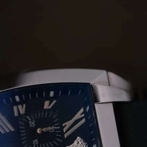 Close-up of the Versace Character Tonneau Swiss Made Chronograph, featuring a rectangular metal case, dark face with Roman numerals, small seconds subdial, and black textured strap against a dark background.