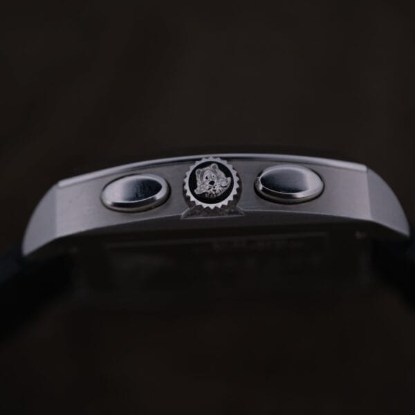 Close-up of the Versace Character Tonneau Swiss Made Chronograph’s side, featuring two round push buttons and a crown with an engraved logo, set against a dark background.