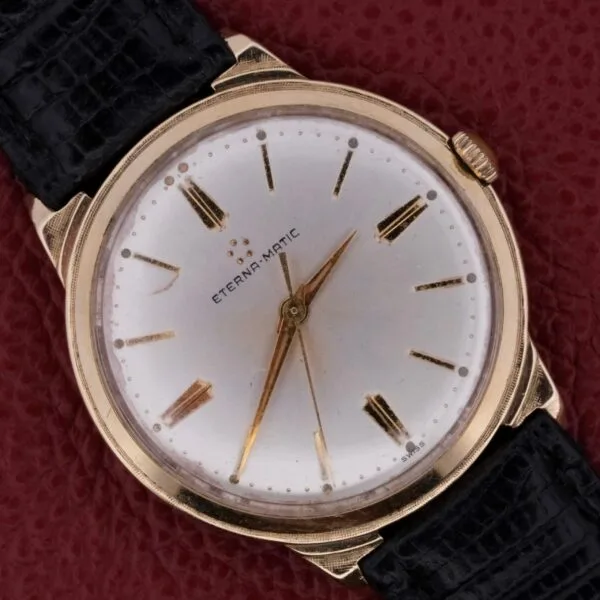 A vintage 14k solid gold Eternamatic mens watch features a sunburst dial with a gold case, white face, and black leather strap. Gold hour markers and hands adorn the dial, with ETERNA-MATIC text and five dots near 12 oclock. The dial shows some wear.