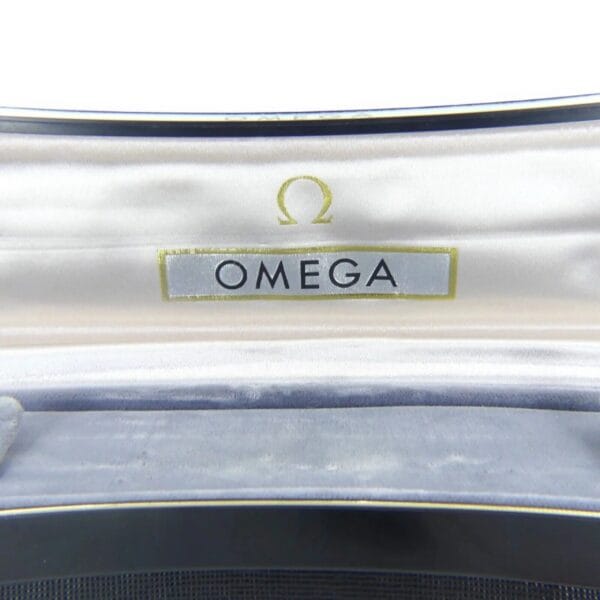 The image reveals the inside lid of a Vintage Omega Continental Can watch box, featuring a white satin lining. The Omega logo, with its iconic symbol and OMEGA text, is printed in black on the fabric within the lid.