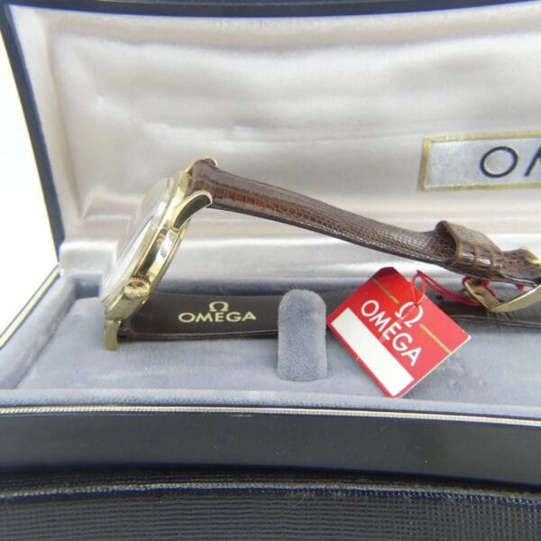 A side view of the gold-colored Vintage Omega Continental Can New Old Stock Men’s Watch with a brown leather strap is presented in a black and gray box. An open lid reveals the brand logo, with a red Omega tag beside the watch.