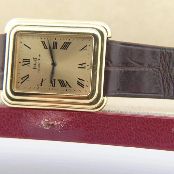 The Vintage Piaget Protocole 74121 features a square, gold-colored design with Roman numerals and black hands on a brown leather strap, set diagonally on a textured red surface.