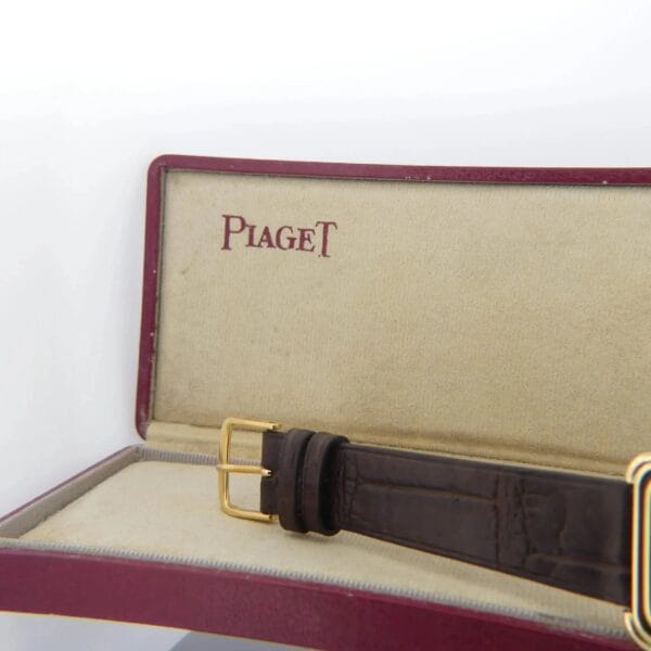 A Piaget Protocole 74121 watch with a brown leather strap and gold buckle is partially visible in a red and beige box with a suede-like interior. The brands name is printed in red on the lid.