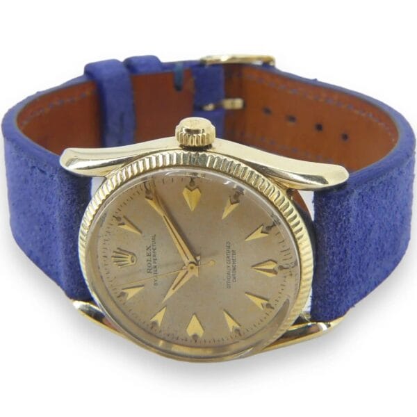 The Vintage Rolex 6593, a 14k Gold Oyster Perpetual with rare Bombay lugs and a fluted bezel, features a light brown dial with gold hour markers. The round-faced watch pairs with a blue suede strap and includes the brand logo on the dial.