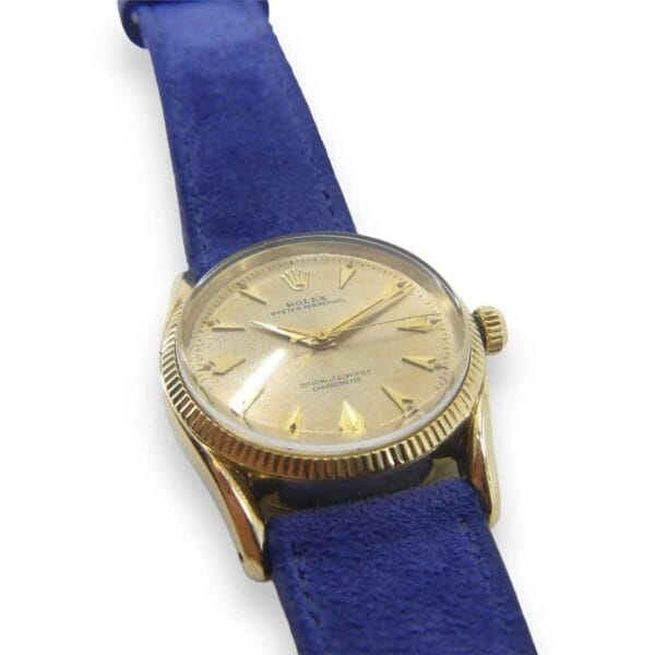 The Vintage Rolex 6593 Oyster Perpetual features a beige dial with triangular markers, brand text, and a crown logo. It includes a blue leather strap attached to gold Bombay lugs, simple watch hands, and a textured fluted bezel.