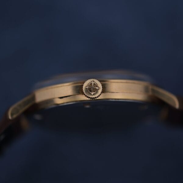 Close-up side view of the Zodiac Hermetic 17 Jewels Dr. Pepper Dial wristwatch case with a textured crown, sharply focused to highlight its fine details against a dark blue background, while the rest of the watch remains slightly blurred.
