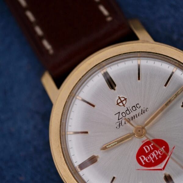A close-up of the Zodiac Hermetic 17 Jewels Dr. Pepper Dial wristwatch with a brown leather strap, gold-tone case, and Dr Pepper logo on a silver face against a textured blue background.