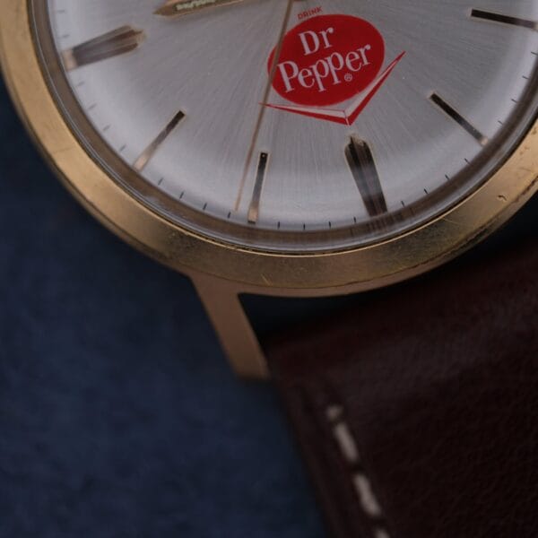 Close-up of the Zodiac Hermetic 17 Jewels Dr. Pepper Dial watch face with gold trim, featuring red Dr Pepper logos in place of numerals, set against a dark blue background and showing part of a brown leather wristband.