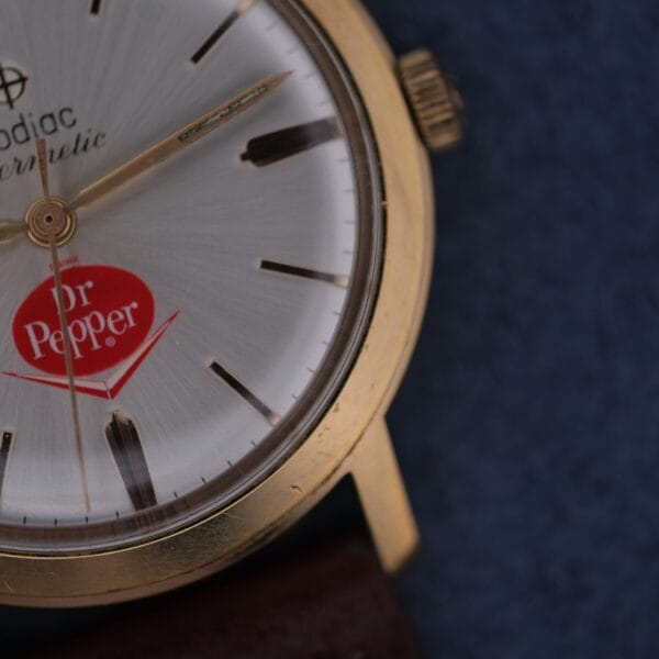 Close-up of the Zodiac Hermetic 17 Jewels watch featuring a gold case, silver dial with black markers, a red Dr Pepper logo at six o’clock, and a brown leather strap.