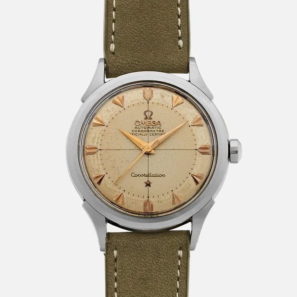 A vintage Omega Costellation 2852 wristwatch with a round beige dial, gold hour markers, and a green leather strap, viewed from the front. The dial displays the Omega logo and “Automatic Chronometer Officially Certified.”.