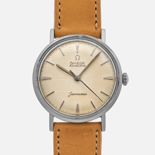 A vintage Omega Seamaster 14765 wristwatch with a cream-colored dial, silver hour markers and hands, brown leather strap, and the words Omega Automatic and Seamaster on the face.