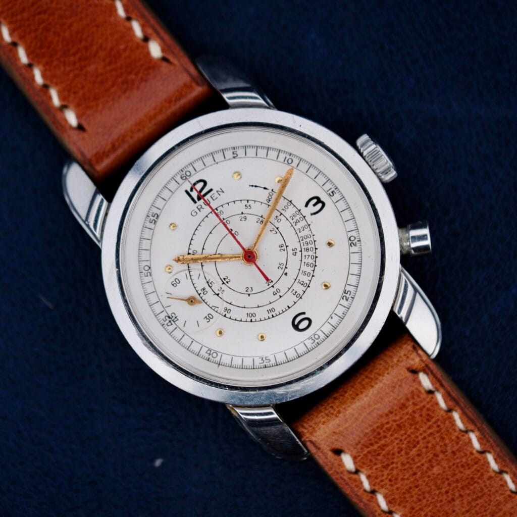 The Gruen Chronotimer Steel With Pulsations Dial features a brown leather strap and white dial with red and gold hands, showcased against a dark background.