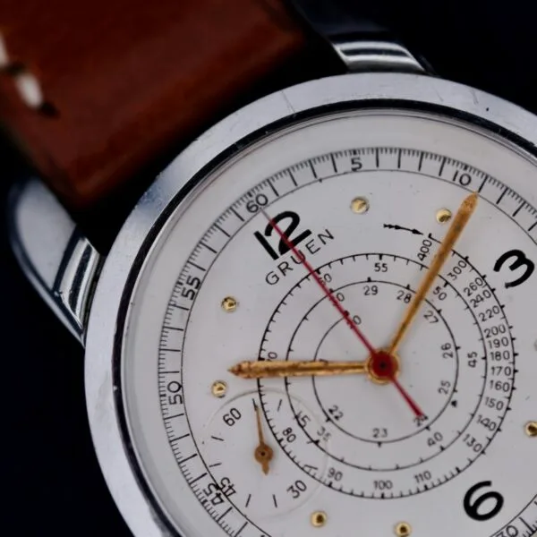 Close-up of the Gruen Chronotimer featuring a white dial, silver casing, and brown leather strap. It has black numerals at 12, 3, 6, and 9, a red central hand, golden subdials, and displays Gruen on the face.