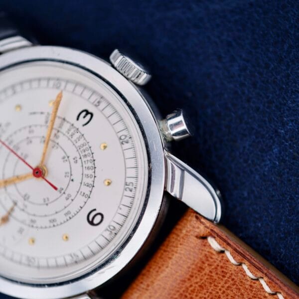 Close-up of a Gruen Chronotimer Steel with Pulsations Dial, featuring a silver watch face with multiple dials and numbers 3 and 6. It has a brown leather strap, small crown, and pusher on the right, set against a dark blue fabric background.
