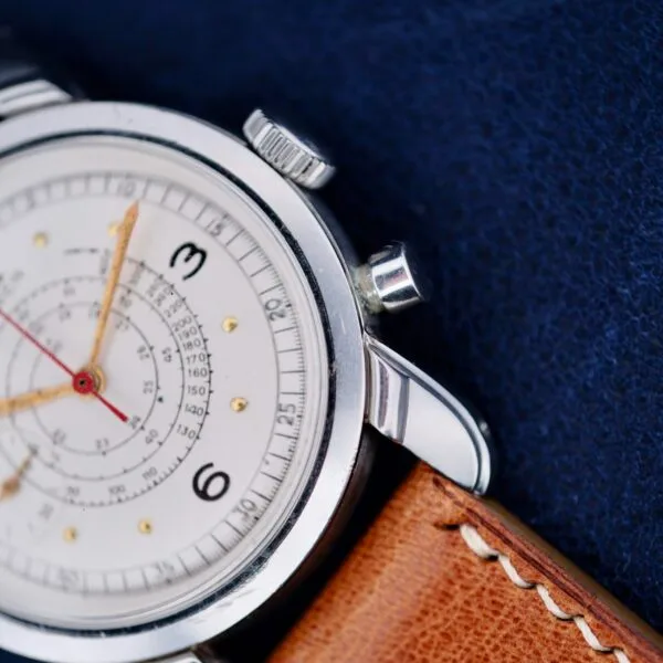 Close-up of a Gruen Chronotimer Steel with Pulsations Dial, featuring a silver watch face with multiple dials and numbers 3 and 6. It has a brown leather strap, small crown, and pusher on the right, set against a dark blue fabric background.