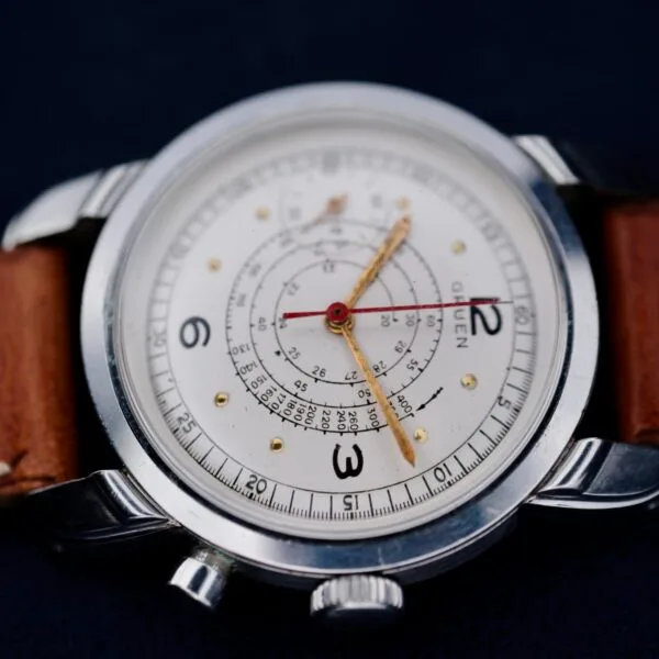 A close-up of the Gruen Chronotimer Steel wristwatch on a dark background, showcasing its brown leather strap, white face with black numbers, gold dot markers, and red hands. The intricate central design includes multiple scales and markings for pulsations.