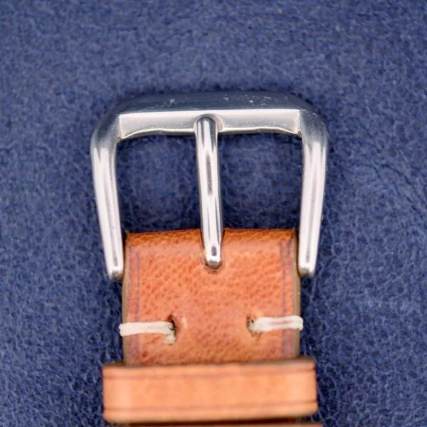Close-up image of a Gruen Chronotimer Steel with Pulsations Dial showing its silver metal belt buckle attached to a brown leather belt stitched with white thread, set against a textured blue fabric background.