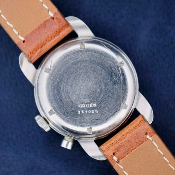 A silver Gruen Chronotimer Steel wristwatch with a leather strap, engraved with 441085, rests on a dark blue surface.