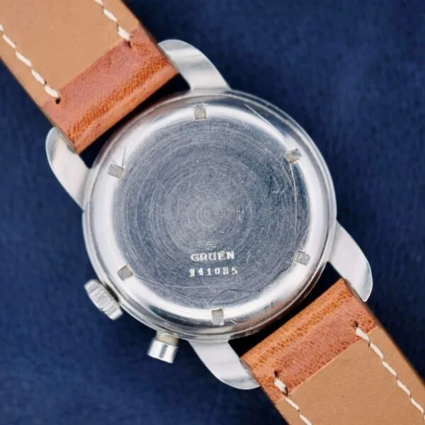 A silver Gruen Chronotimer Steel wristwatch with a leather strap, engraved with 441085, rests on a dark blue surface.