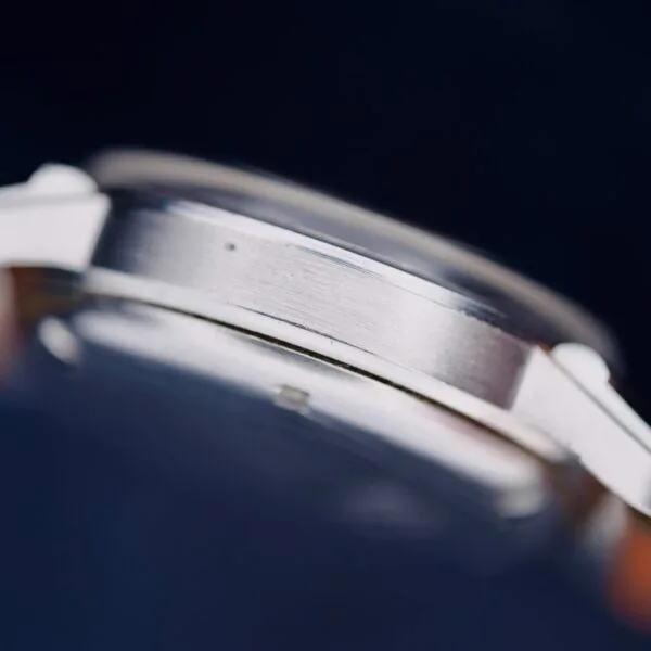 Close-up of the side profile of a Gruen Chronotimer Steel wristwatch, highlighting its brushed metal texture. The focus is on the watchs edge against a dark, blurred background, with the face and other details out of frame.