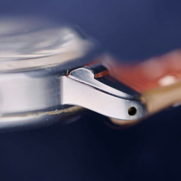 A close-up of the Gruen Chronotimer Steel watch case highlights a silver metal lug with a hole at the tip, featuring part of the crystal and a copper-colored strap against a dark blue backdrop.