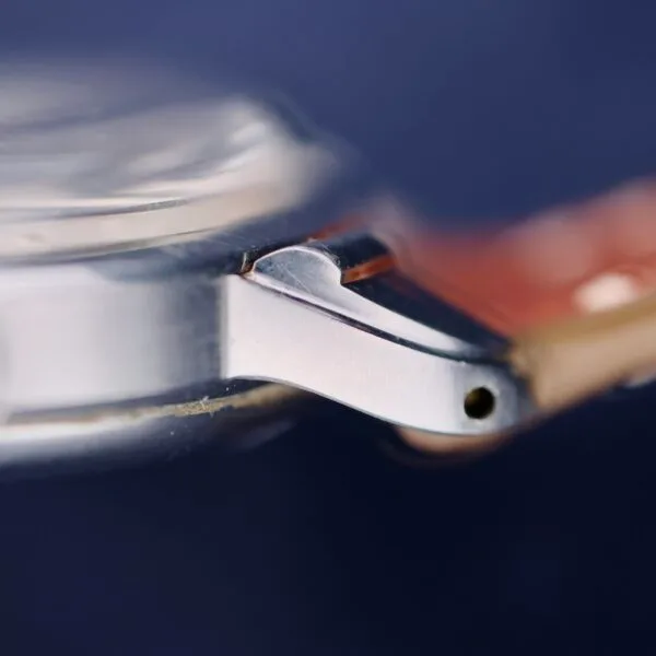 A close-up of the Gruen Chronotimer Steel watch case highlights a silver metal lug with a hole at the tip, featuring part of the crystal and a copper-colored strap against a dark blue backdrop.
