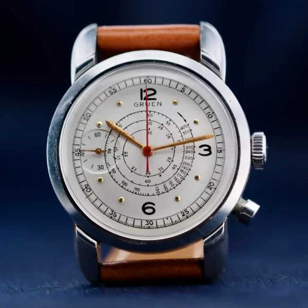 A close-up of the Gruen Chronotimer Steel features a silver case and brown leather strap, a white dial with a central red second hand, black numerals at 12, 3, 6, and 9, an inner ring with markings, and a right-side crown.
