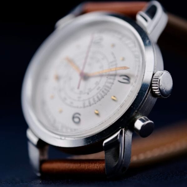 Close-up of the Gruen Chronotimer Steel with Pulsations Dial, featuring a silver casing and brown leather strap. The white watch face displays black numerals and multiple sub-dials, with the time set to about 10:08, focusing on the side and crown.