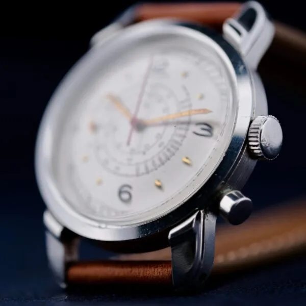 Close-up of the Gruen Chronotimer Steel with Pulsations Dial, featuring a silver casing and brown leather strap. The white watch face displays black numerals and multiple sub-dials, with the time set to about 10:08, focusing on the side and crown.