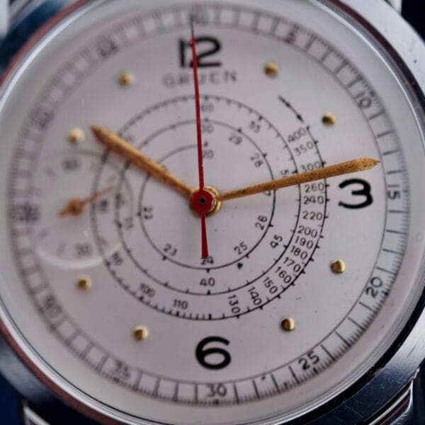 Close-up of a Gruen Chronotimer Steel watch face, featuring gold-tone hour and minute hands, a red second hand, black numbers at 12, 3, 6, and 9, concentric circular scales marked in black. GREEN is below the 12.