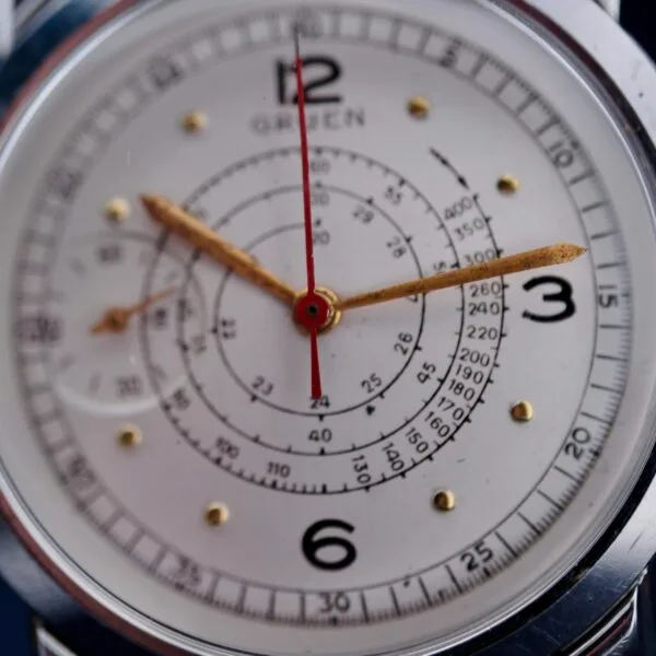Close-up of a Gruen Chronotimer Steel watch face, featuring gold-tone hour and minute hands, a red second hand, black numbers at 12, 3, 6, and 9, concentric circular scales marked in black. GREEN is below the 12.